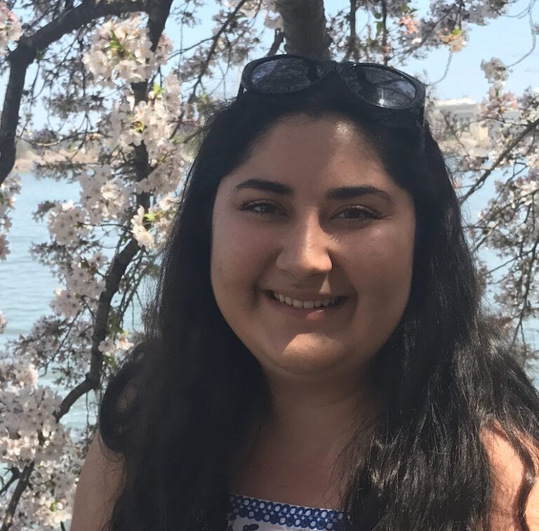 A woman with dark hair and sunglasses on her head smiling with a background of tree branches with white blossoms.