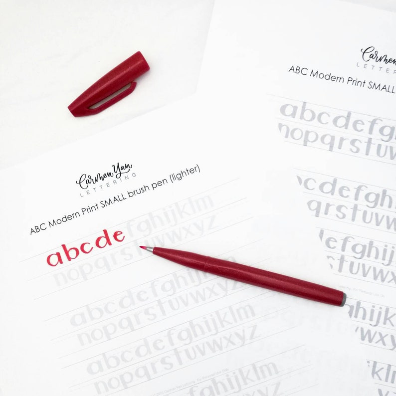 May include: A close-up of a white sheet of paper with gray printed letters of the alphabet. The letters are in a modern print font. A red brush pen is lying on the paper, and the letters "a b c d e" are written in red ink.