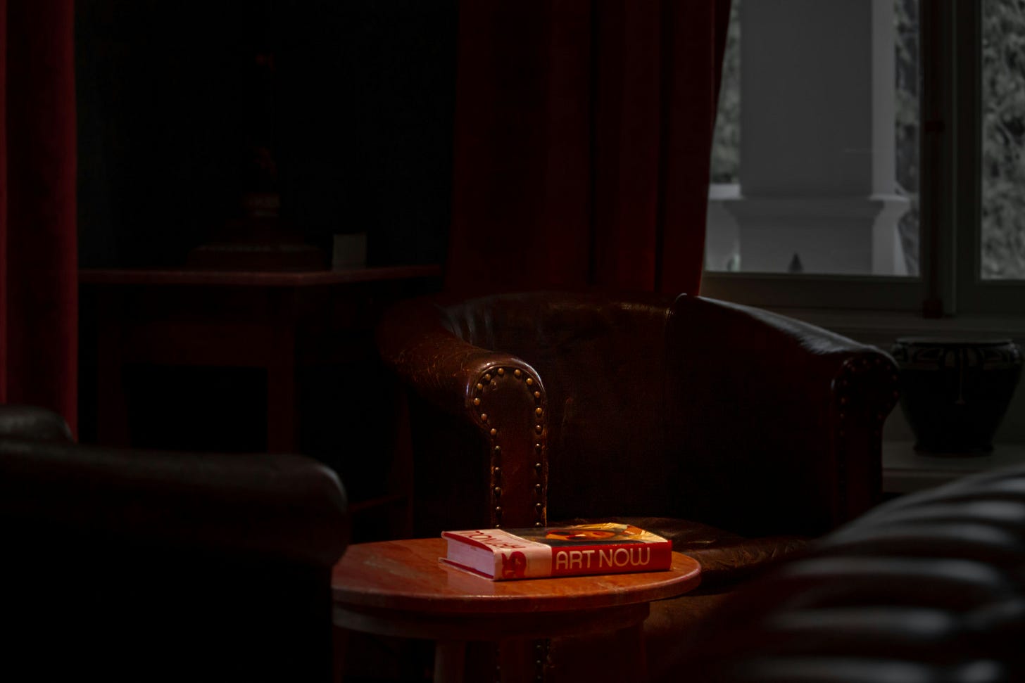 A moody, low-light photograph of an empty, classic leather armchair tucked into a shadowed corner. In the foreground, a small wooden side table holds a book. In the background, a large window looks out onto a soft grey daylight, framed by deep red curtains.