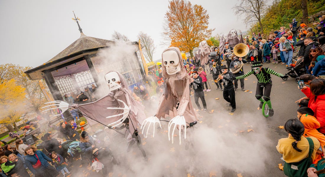 A Halloween parade with skeleton puppets, a marching band, and crowds of people in Halloween outfits