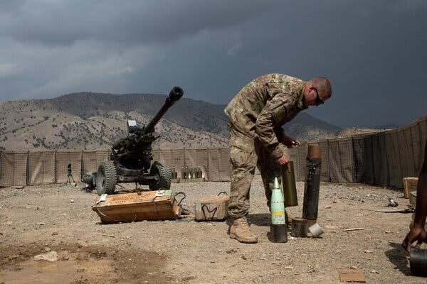 A person in camouflage gear leans over a blue-green projectile in a dusty landscape near a large, two-wheeled weapon.