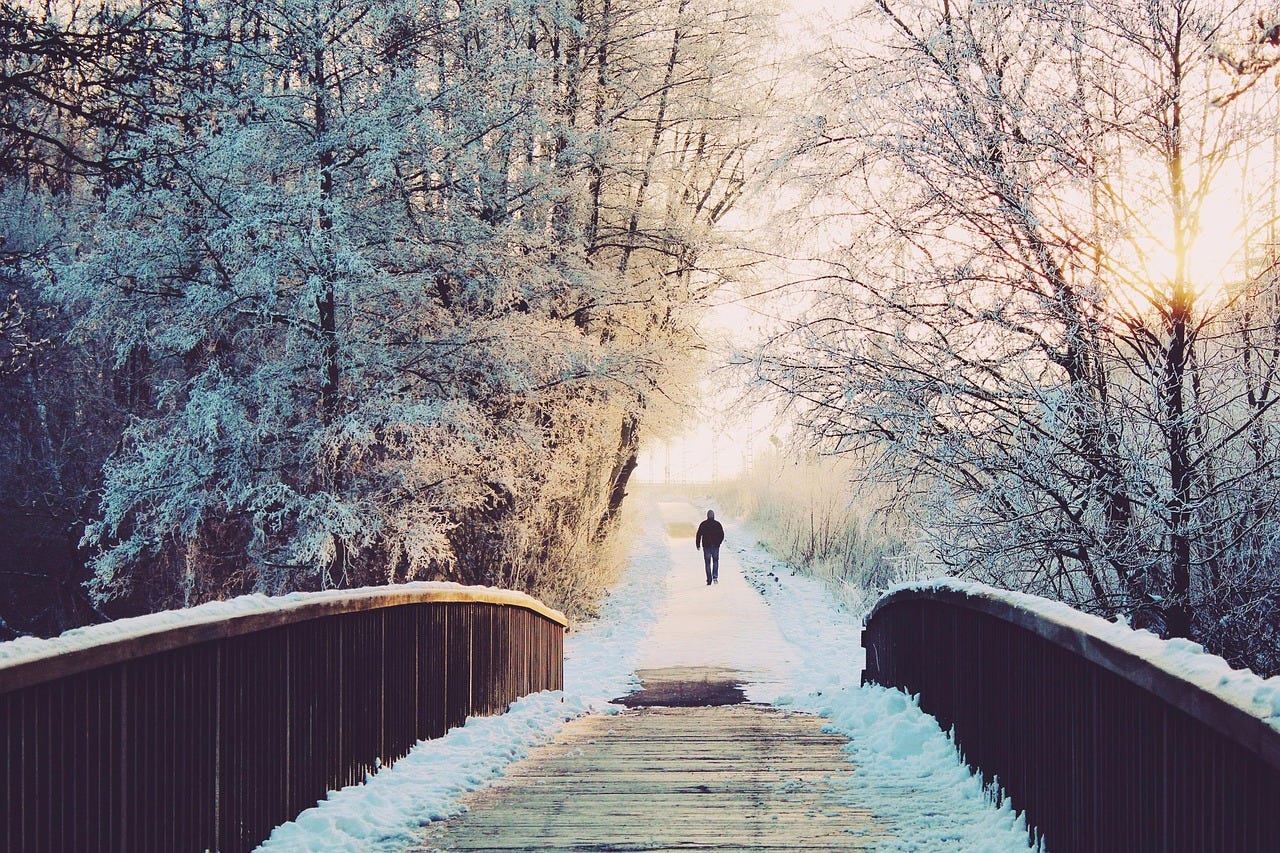 A long path covered in snow, a wooden bridge where the back of a man can be seen walking in the distance. A long path covered in snow, a wooden bridge where the back of a man can be seen walking in the distance.
