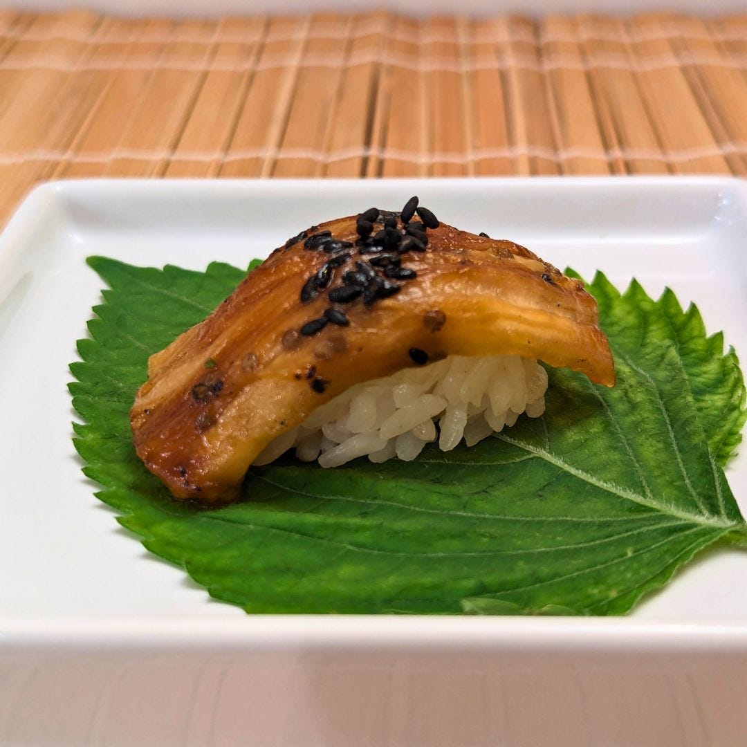 Plant-based eggplant unagi nigiri on a shiso leaf with black sesame garnish, plated simply on white.
