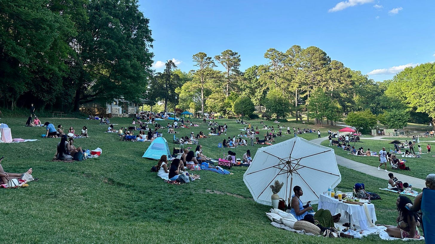 Sunny park with many families picnicking on a grassy lawn near trees. Tents and a white canopy are in the foreground; groups sit on blankets under a clear blue sky with a few clouds.