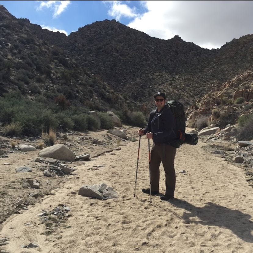 Image: A man with a backpacking pack and hiking polls stands in the middle of a sandy path in the desert infront of a mountain range.