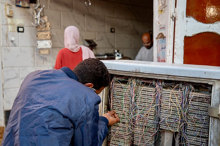 Top left: A busy market street in Aswan with vendors and shoppers beneath a mosque minaret and tangled overhead wires Top right: Two women sitting together sharing a phone beside a table of cosmetics and bottled goods, with a woman in a niqab seated nearby Bottom right: An elevated view across the rooftops of old Aswan toward the Nile, with feluccas and cruise ships moored along the banks and the modern city beyond Bottom left: A young man crouched over an open telecommunications junction box with tangled colored wires, while a woman in a pink hijab and a shopkeeper stand in the doorway behind him
