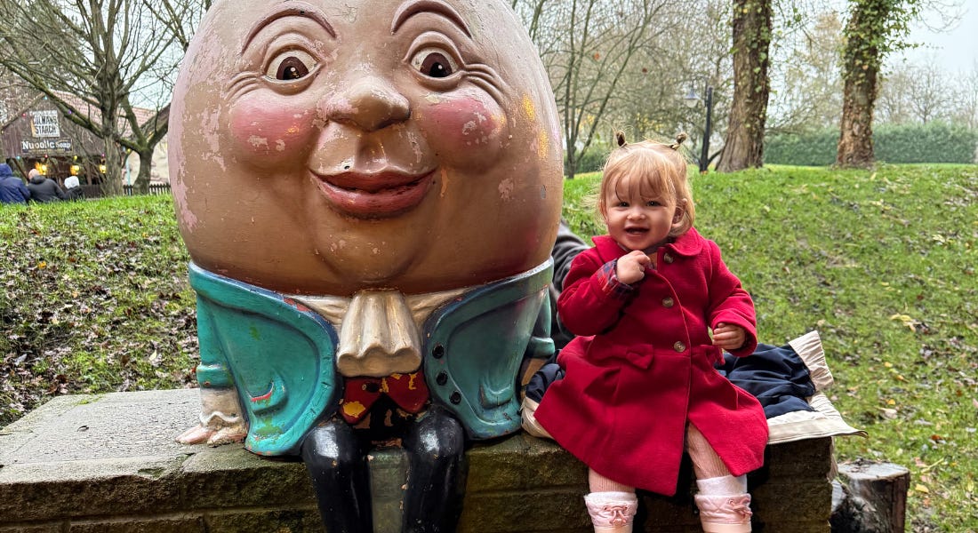 A young girl in a red coat sat next to a Humpty Dumpty statue