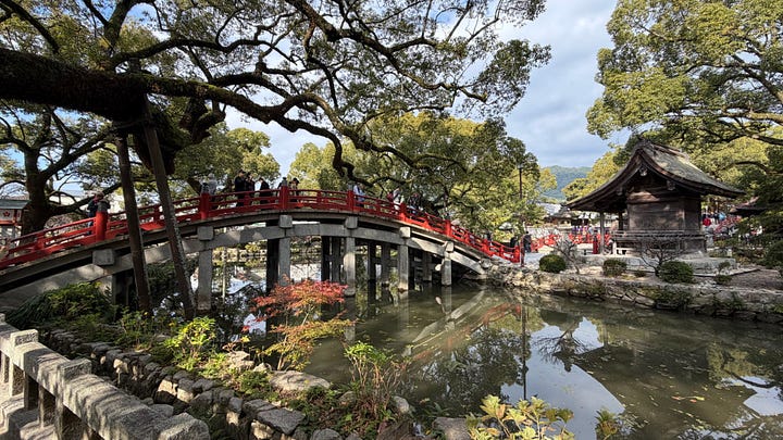 Image 1 alt text: A traditional Japanese dining room with dark wood tables and chairs, framed artwork on the walls, and large windows opening onto a leafy garden courtyard.  Image 2 alt text: A scenic shrine garden with a curved red bridge crossing a reflective pond, surrounded by trees and traditional wooden buildings.  Image 3 alt text: An accessible bathroom with a roll-in shower, bathtub, grab bars, handheld shower, raised toilet, sink with clearance underneath, and a blue shower chair.  Image 4 alt text: A modern hotel room with two twin beds, soft lighting, floor-to-ceiling curtains, and a small sitting area with a round table and armchair.