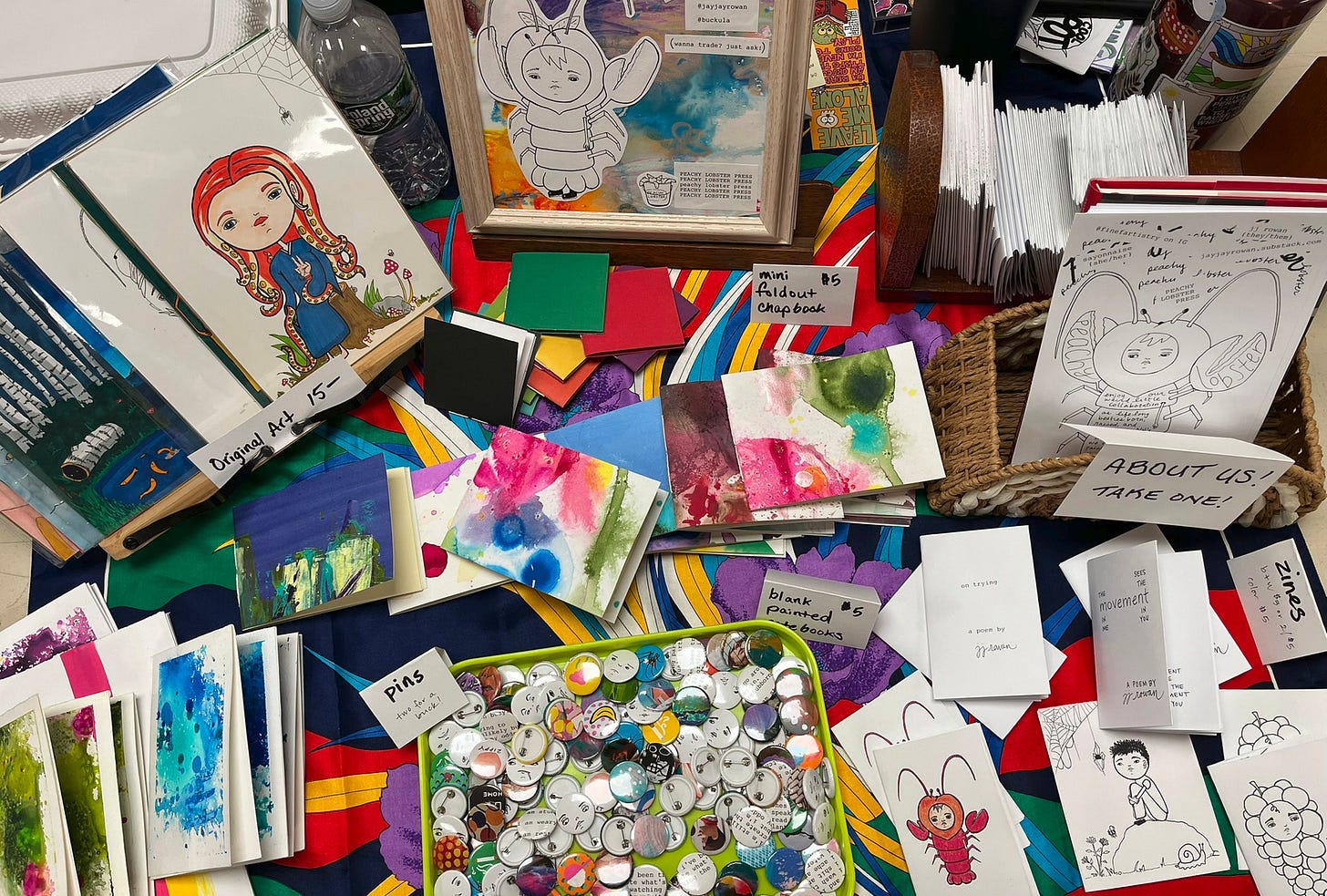 a zine fest table full of colorful pins, zines, painted notebooks, illustrations, and other ephemera