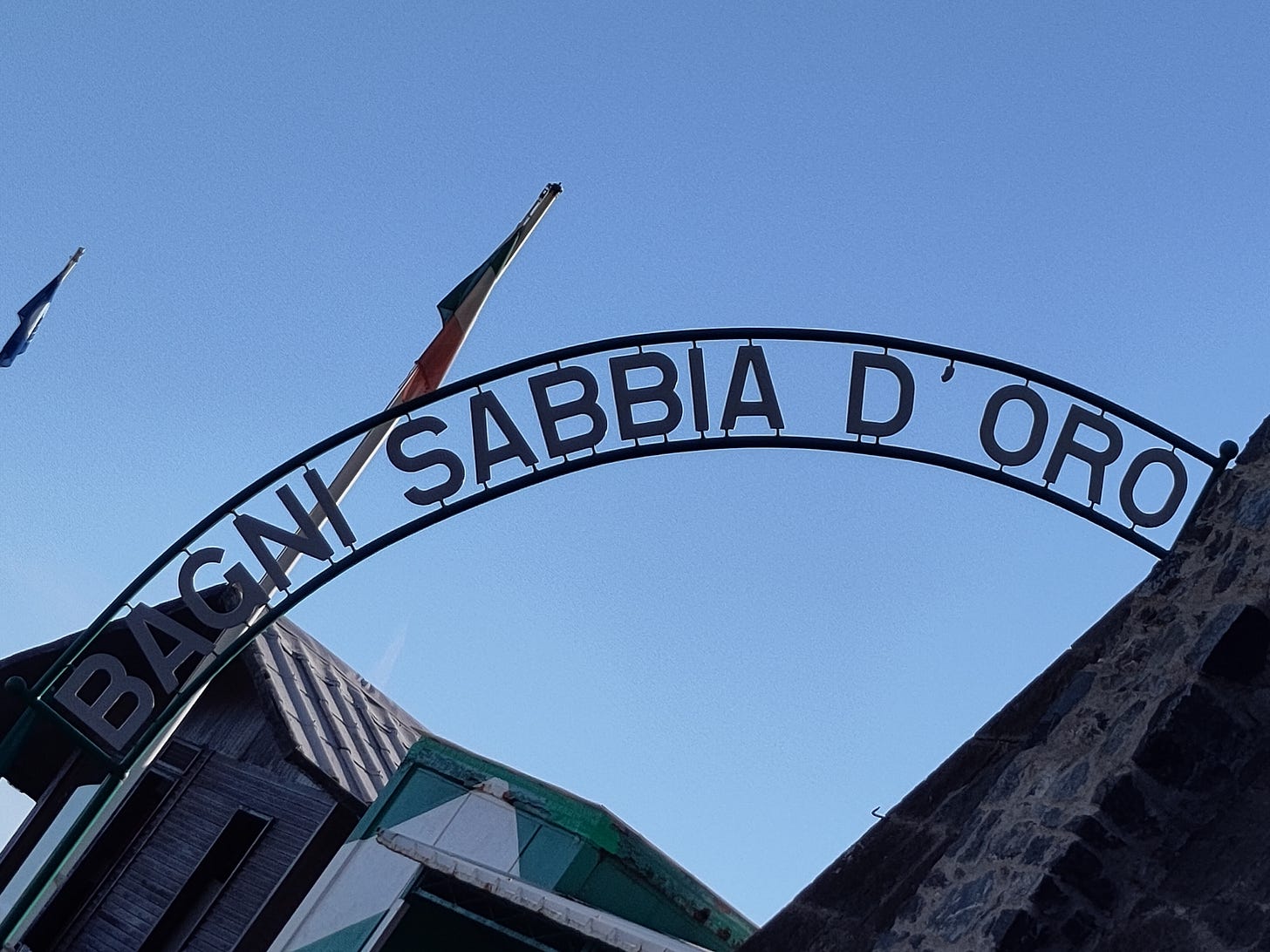 Fotografia dove in primo piano si trova un'insegna su cui si legge "Bagni Sabbia D'Oro" e, in secondo piano, si vedono delle parti di edifici in muratura e legno e delle bandiere. Sullo sfondo c'è un cielo blu privo di nuvole.