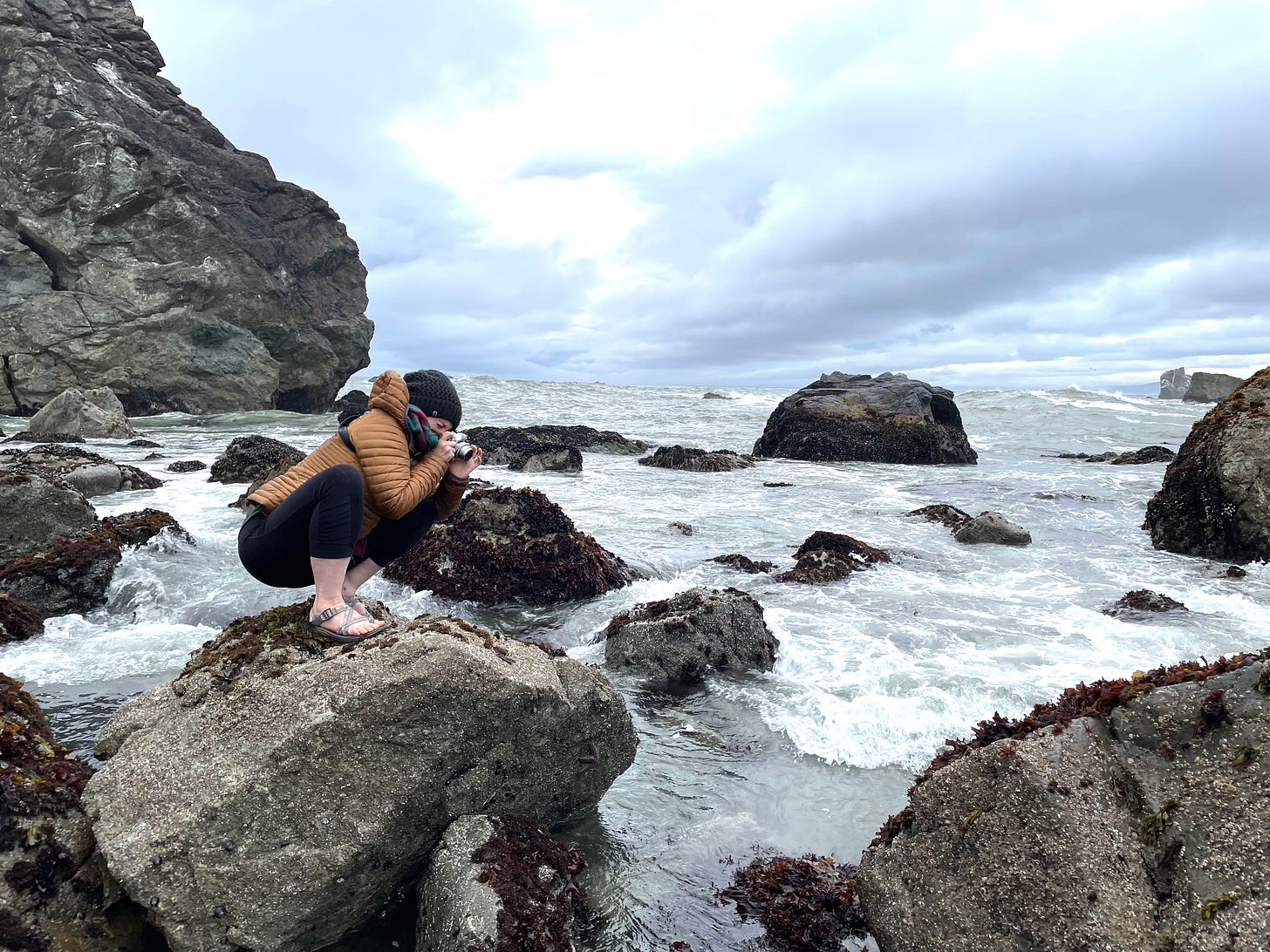 Photo of Pam crouched down on a boulder with her camera taking photos of starfish in tidal pools. Small ocean waves and large boulders surround her under a cloudy sky. Pam is wearing Chaco sandals, black pants and a warm coat and hat.