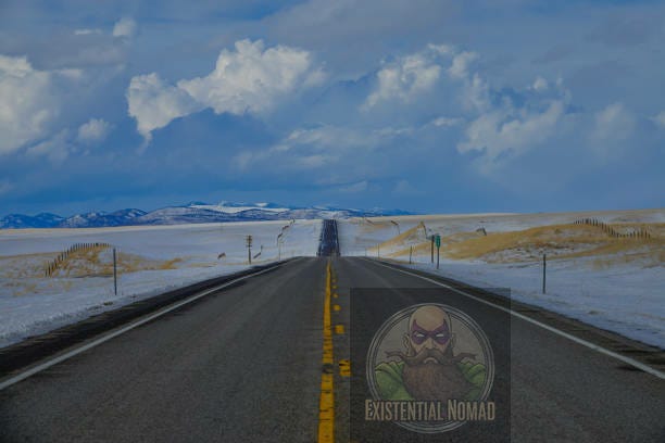 This is a photograph of a long, straight road stretching toward the horizon, with a double yellow line running down the middle. The landscape on either side of the road is covered in snow and dry, golden grass. In the far distance, snow-capped mountains are visible under a wide, cloudy blue sky. The scene has a cold, expansive, and somewhat desolate feel.