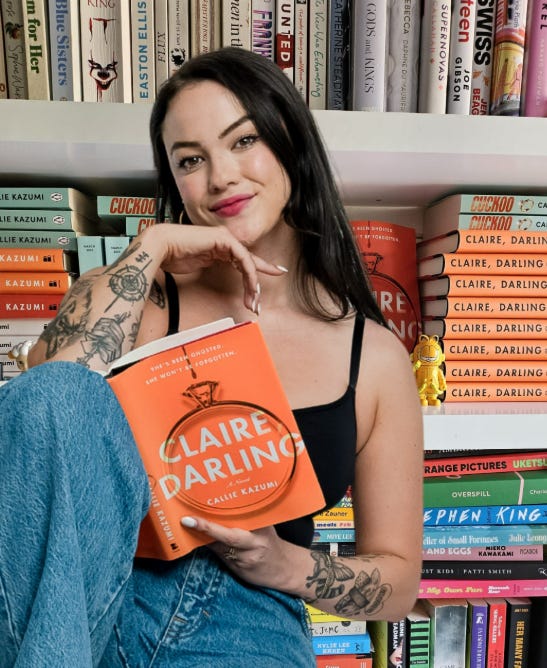 Well-lit image of author smiling, holding her own book with the cover and title facing outwards, in front of a bright and packed bookshelf