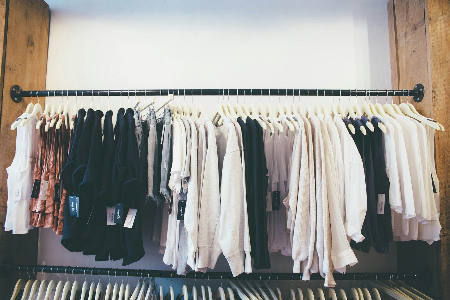 A full clothing rail with neutral-toned garments on white hangers, no sale or discount tags visible, in a warm, naturally lit retail space with wooden wall panels.