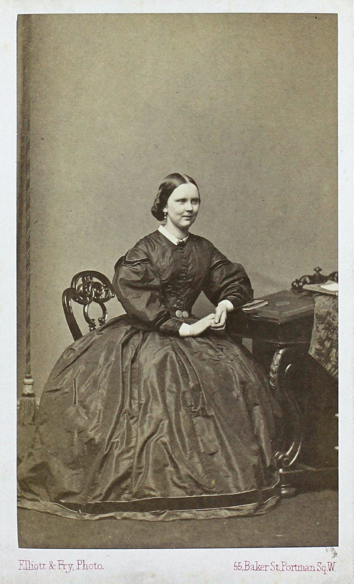 An old photo of a woman sat at a desk, posing with her hands clasped in front of her
