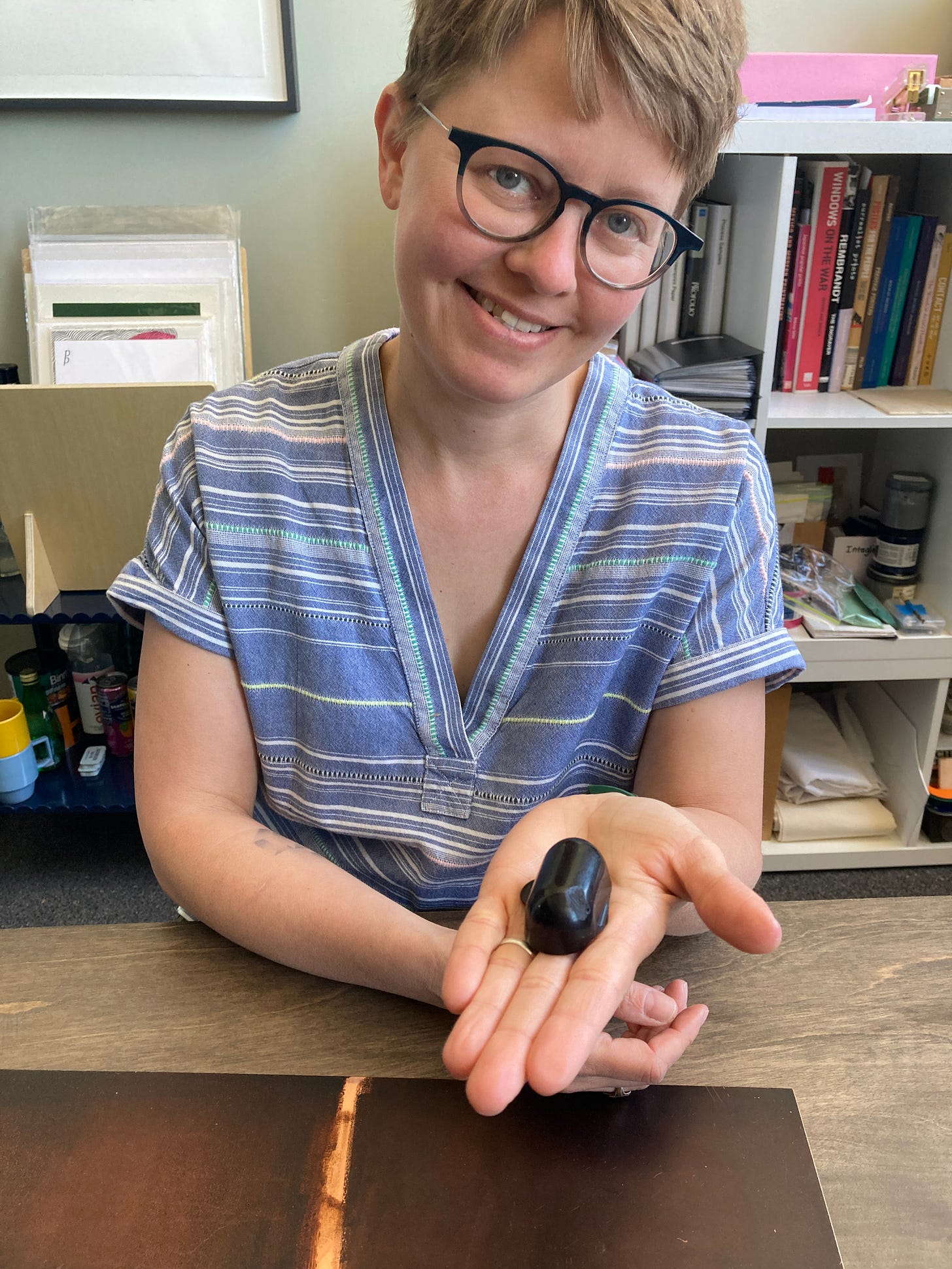 A white woman with short blonde hair and glasses holds a black ball of ground in the palm of her hand. She is smiling and looking at the camera. 