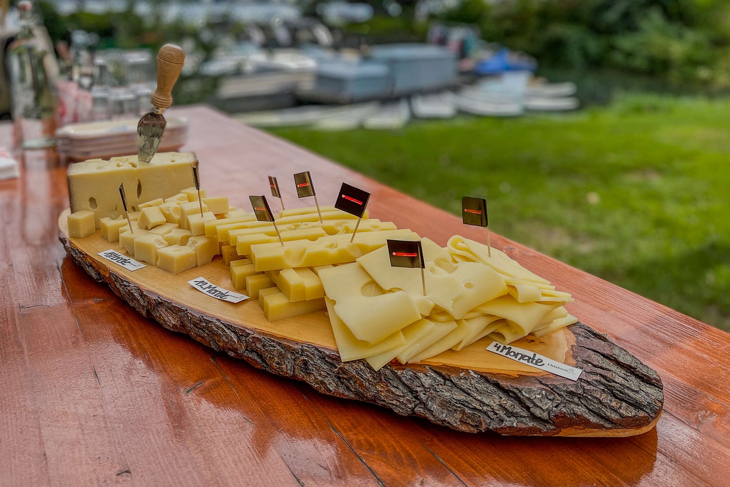 Holzbrett mit Rindenrand, darauf Emmentaler AOP in verschiedenen Reifegraden angerichtet: von milden Würfeln über mittelreife Stücke bis zu Scheiben des 14 Monate gereiften Käses. Kleine Fähnchen und Beschriftungen markieren die Unterschiede, im Hintergrund unscharf eine Wiese und Boote am Wasser. Holzbrett mit Rindenrand, darauf Emmentaler AOP in verschiedenen Reifegraden angerichtet: von milden Würfeln über mittelreife Stücke bis zu Scheiben des 14 Monate gereiften Käses. Kleine Fähnchen und Beschriftungen markieren die Unterschiede, im Hintergrund unscharf eine Wiese und Boote am Wasser.