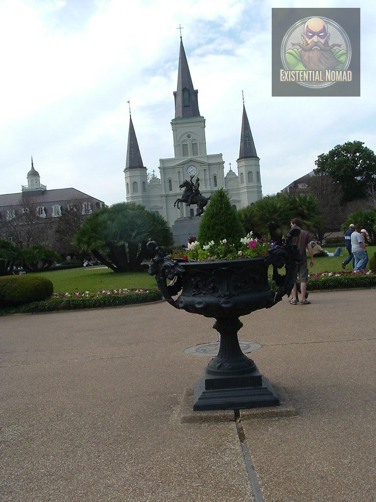 This is a photograph taken in Jackson Square, New Orleans. In the foreground, there is a large, ornate, dark-colored planter filled with flowers and greenery. In the background, the iconic St. Louis Cathedral is visible with its three spires and a statue of Andrew Jackson on a horse in front of it. The sky is partly cloudy, and people are walking and relaxing on the grass and pathways of the square.