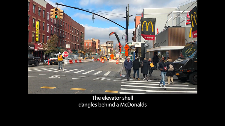 A photo of an intersection in a city, with a worker directing pedestrian traffic and a McDonalds on a corner. Beneath the photo, against a black background, there’s a caption that says, The elevator shell dangles behind a McDonalds.