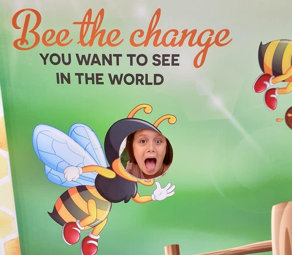 a picture board that says, bee the change you want to see in the world. There is a painting of a bee, with a space for a child to put their head through. A child is looking out with an animated face.
