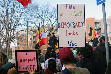 Left image: one small square pink poster reading "I support unions" and a person in a red cap holds up a poster in rainbow lettering "this is what DEMOCRACY looks like." Middle image: A young Brown man stands to the left of the image facing right holding a microphone and cellphone, a white person with short brown hair and a black shirt stands looking downward in the right corner. Right image: a white poster with black text that reads "The working class holds the power" and a red flag with the letters FRSO (Freedom Road Social Organization) flies in the background
