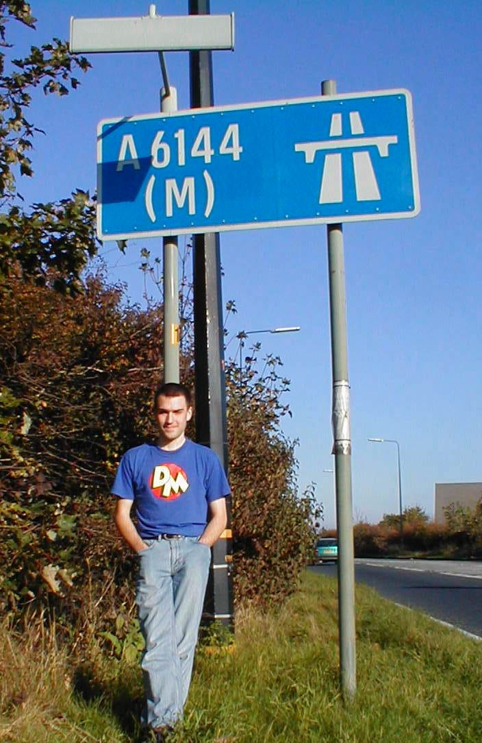 Photograph of a young man in a Danger Mouse t-shirt - that would be your author - striking a casual pose beneath a large blue motorway sign bearing the number "A6144(M)" Photograph of a young man in a Danger Mouse t-shirt - that would be your author - striking a casual pose beneath a large blue motorway sign bearing the number "A6144(M)"
