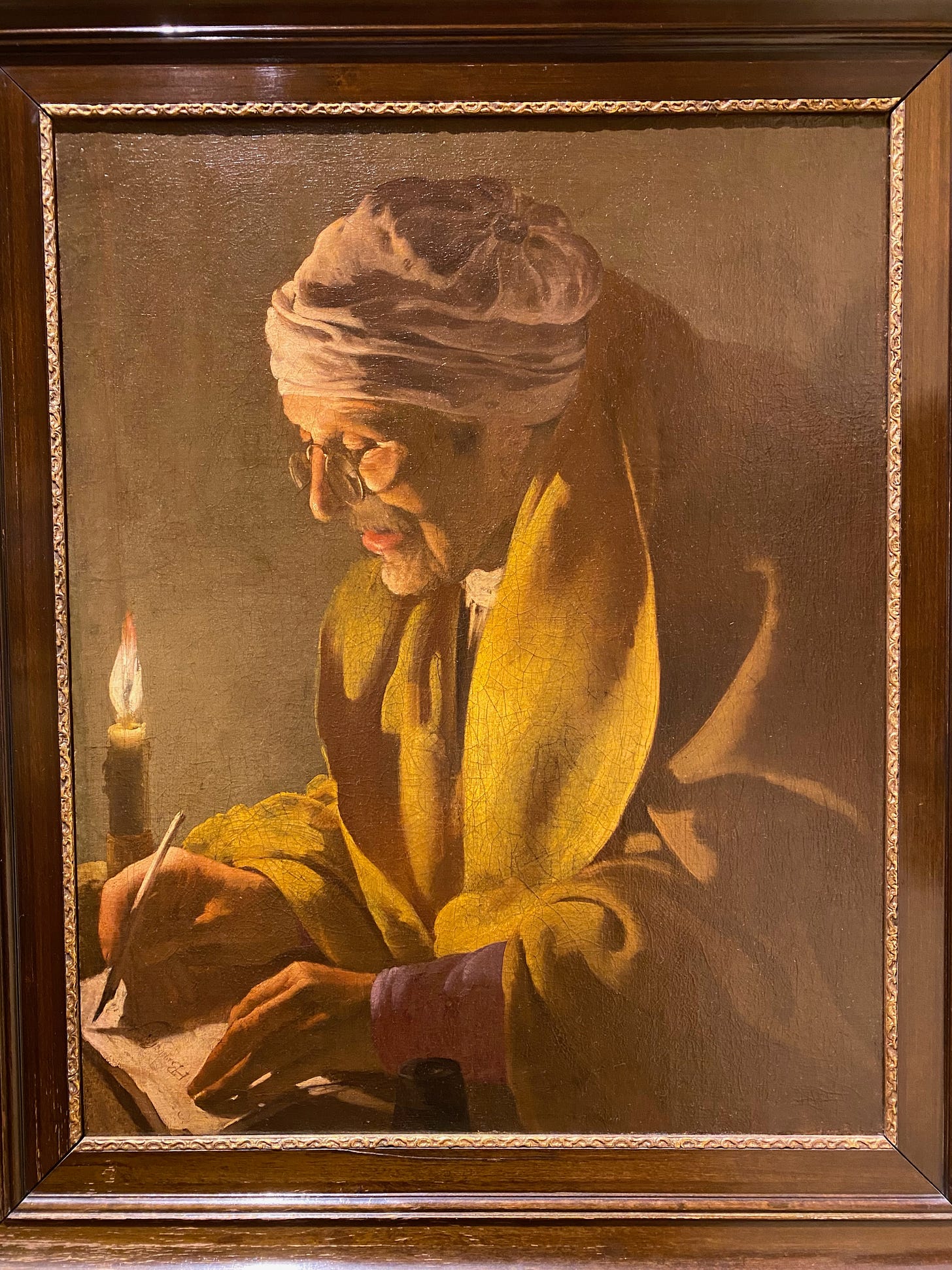 Oil painting of an old man in a white cap and dressing gown writing at a small desk by candlelight.