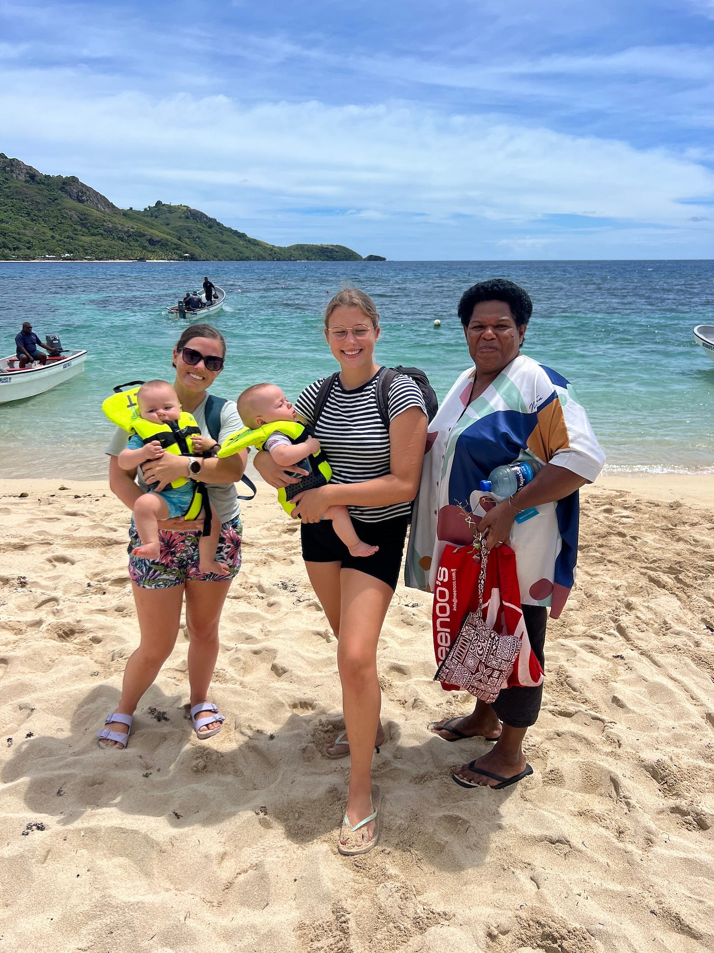 Three women standing on a beach, two of them holding babies with life preservers around their necks. 