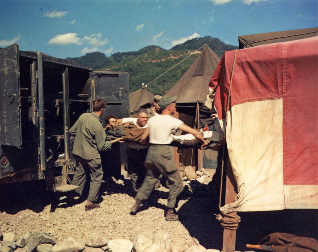 Several uniformed men carry a stretcher with a patient from a military vehicle toward a medical tent with a red cross symbol in a rocky outdoor setting.