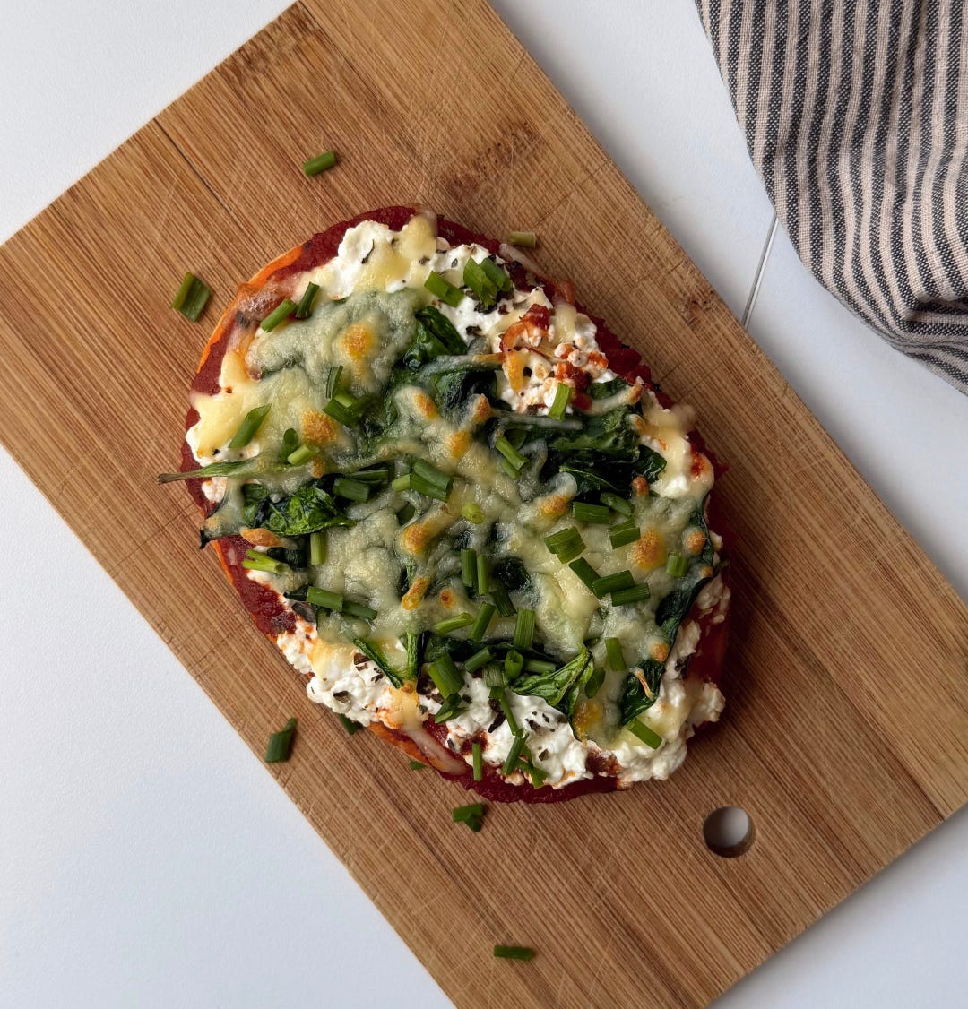 Overhead shot of a high-protein cheesy flatbread bake with melted mozzarella, fresh spinach, and cottage cheese, garnished with chopped chives on a plate. Overhead shot of a high-protein cheesy flatbread bake with melted mozzarella, fresh spinach, and cottage cheese, garnished with chopped chives on a plate.