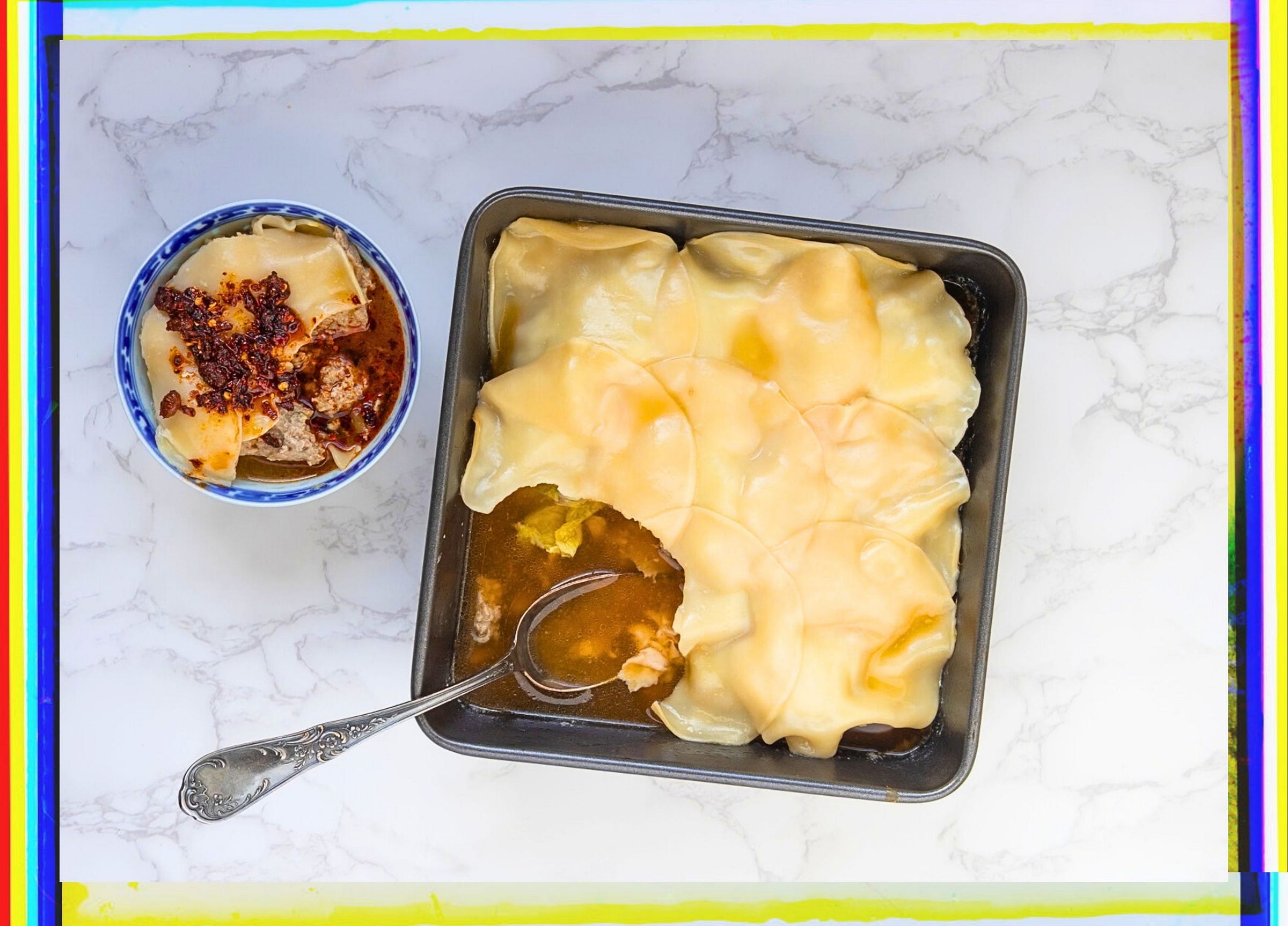 Overhead view of a soup dumpling casserole in a square dark pan with a spoon breaking through the dumpling wrapper top layer, revealing golden broth beneath. A small blue and white bowl with dumplings topped with chili crisp sits alongside on a white marble surface.