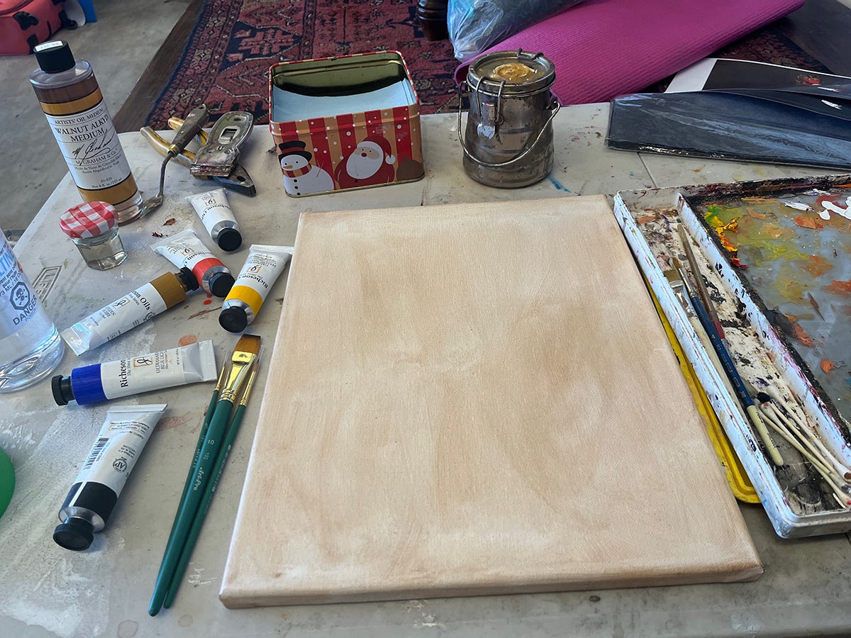 View of a table top with a mixture of oil painting supplies including an assortment of paints, brushes and a canvas that's been brushed with a light coating of brown paint.