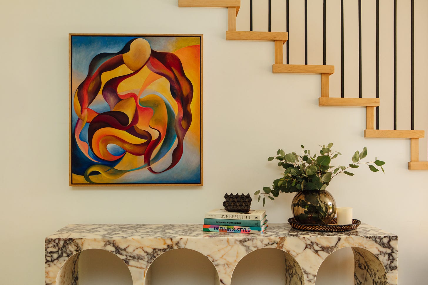 Entryway with Alex Maceda's two bodies in orbit, a painting of warm red, ochre and gold forms against blue, framed in light oak on a cream wall beneath an open oak staircase, above a marble console with books and eucalyptus in a glass vase.