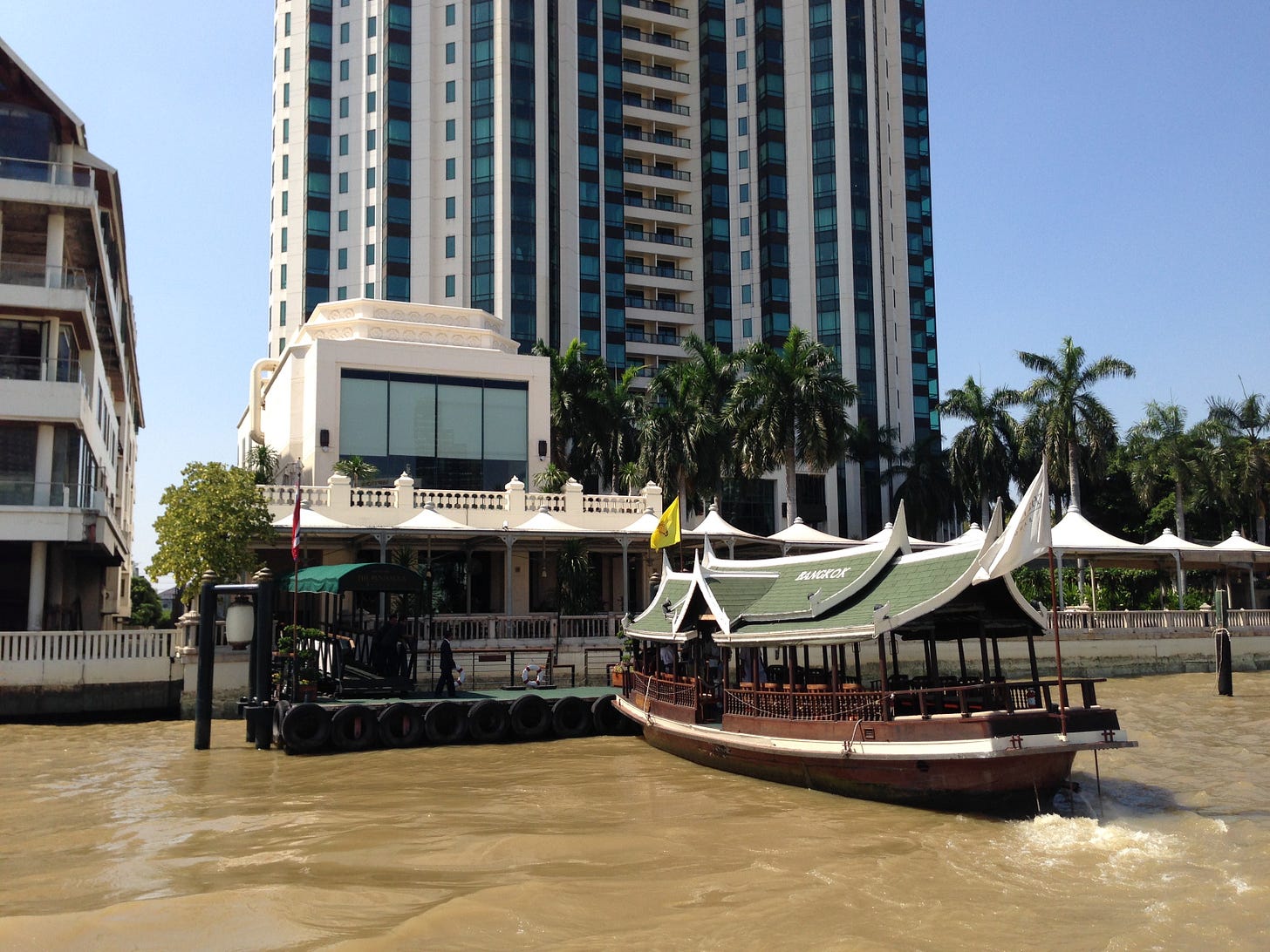 A river boat on the Chao Phraya River in Bangkok, moored beside a modern high-rise hotel and traditional riverside buildings, with palm trees lining the waterfront under a clear blue sky. A river boat on the Chao Phraya River in Bangkok, moored beside a modern high-rise hotel and traditional riverside buildings, with palm trees lining the waterfront under a clear blue sky.