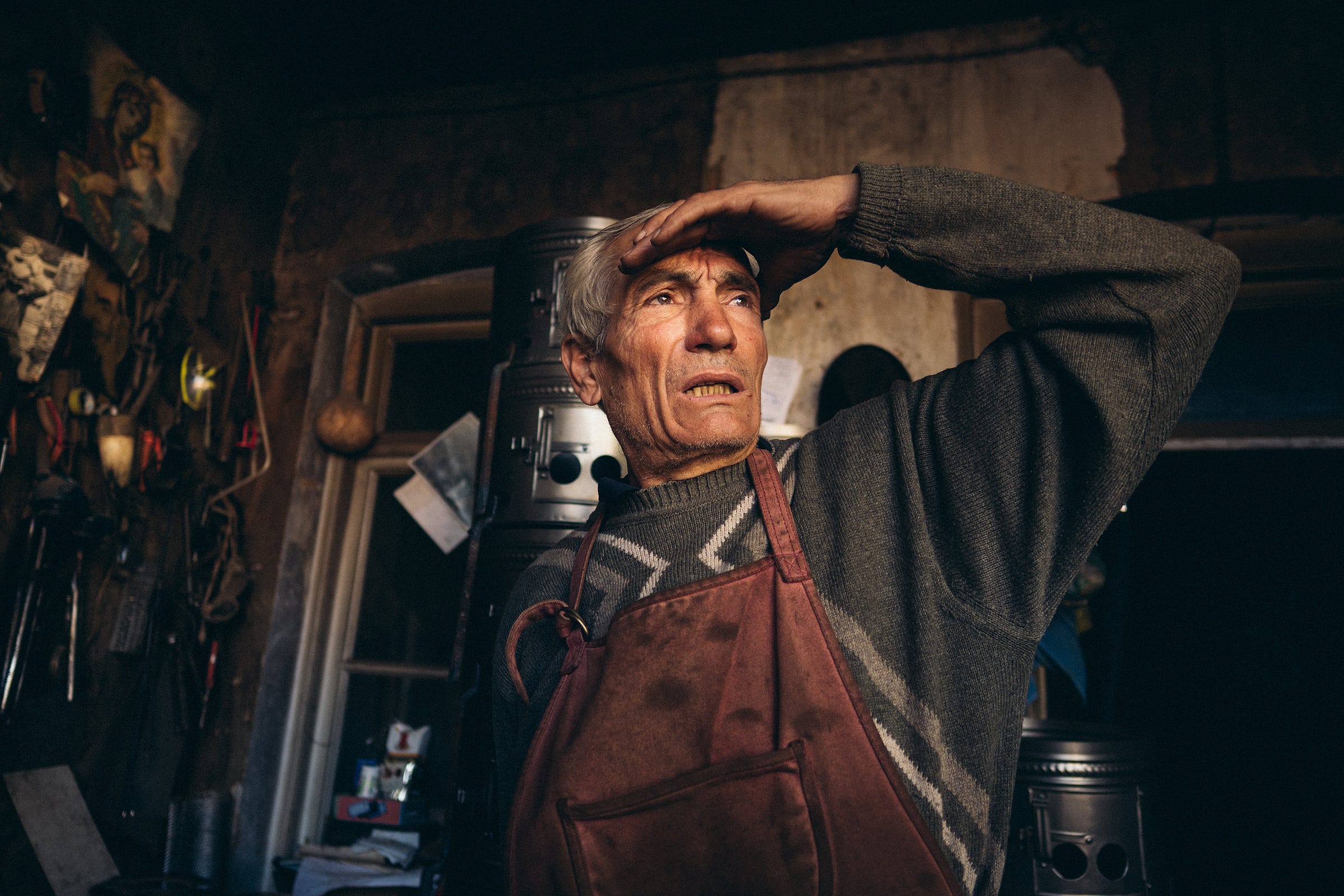 An older man wearing a sweater and apron stands indoors, shading his eyes with one hand as he looks into the distance. The background is cluttered with tools and dimly lit, giving a rustic, workshop-like atmosphere.