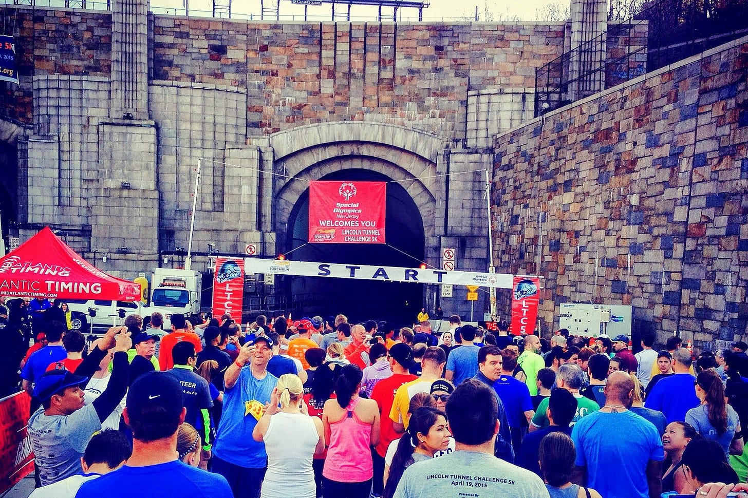 The author stands in a crowd of runners at the start line of the Lincoln Tunnel Challenge 5K. A red Special Olympics banner hangs above the stone tunnel entrance, and a white START banner stretches across the road. Runners in colorful athletic wear wait to enter the tunnel on an overcast day. The author stands in a crowd of runners at the start line of the Lincoln Tunnel Challenge 5K. A red Special Olympics banner hangs above the stone tunnel entrance, and a white START banner stretches across the road. Runners in colorful athletic wear wait to enter the tunnel on an overcast day.