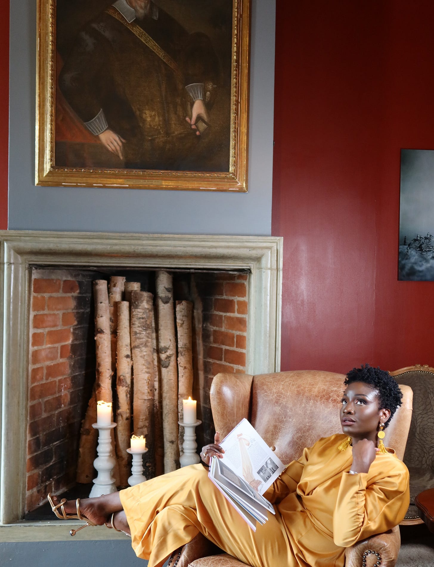 Lovette Jallow in a mustard satin gown sits confidently in a vintage leather armchair, reading a book in front of a fireplace with birch logs and candles. A historical portrait of a white man hangs above the mantle. The setting is regal and defiant.