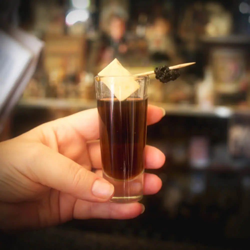 A shot glass filled with an auburn-coloured drink, topped with a cube of white cheese on a stick