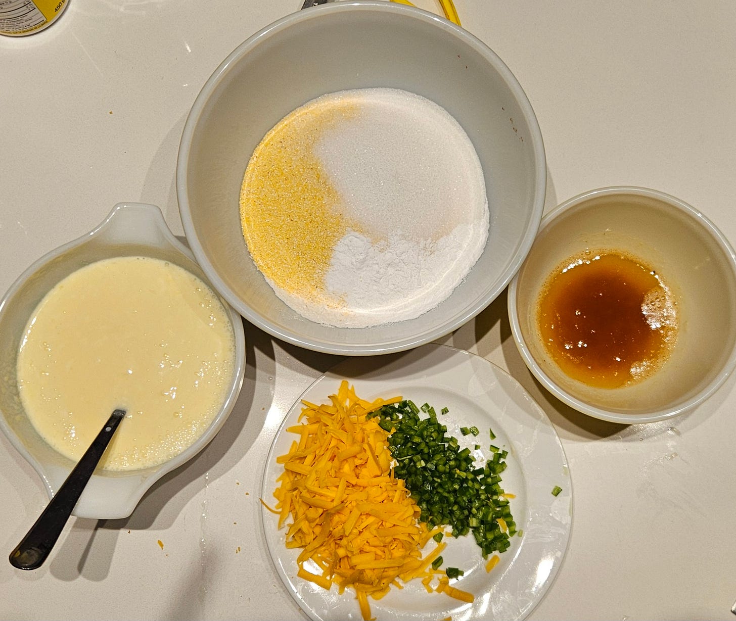 Three bowls. One with wet ingredients, one with dry ingredients, one with browned butter, and a plate with grated cheddar cheese and finely diced jalapeño peppers
