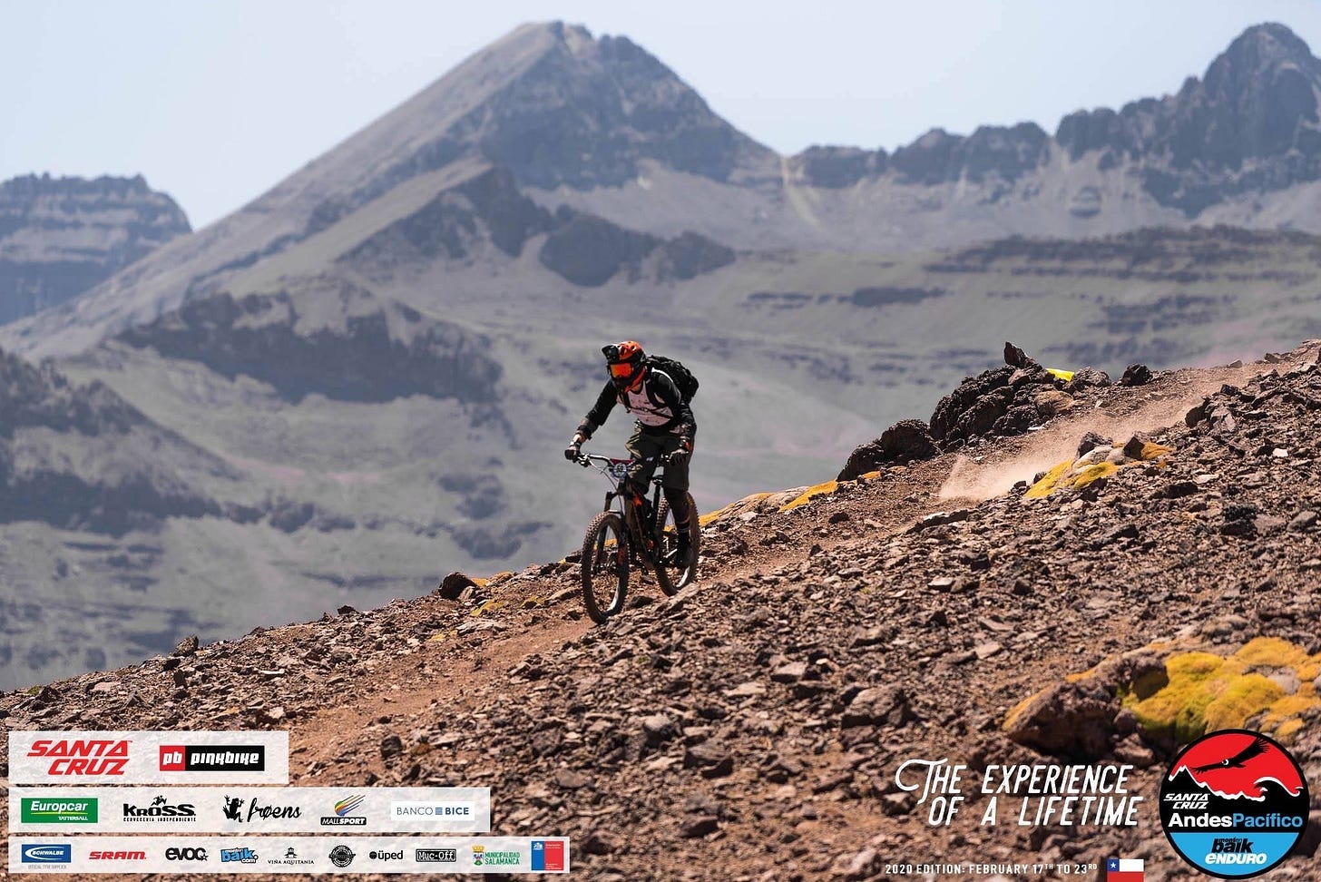A mountain biker wearing a helmet and backpack navigating a steep, rocky mountain trail in the Andes with high peaks in the background.A mountain biker in full gear racing down a steep, rocky mountain ridge during the Andes-Pacifico enduro, with massive, jagged peaks in the background.