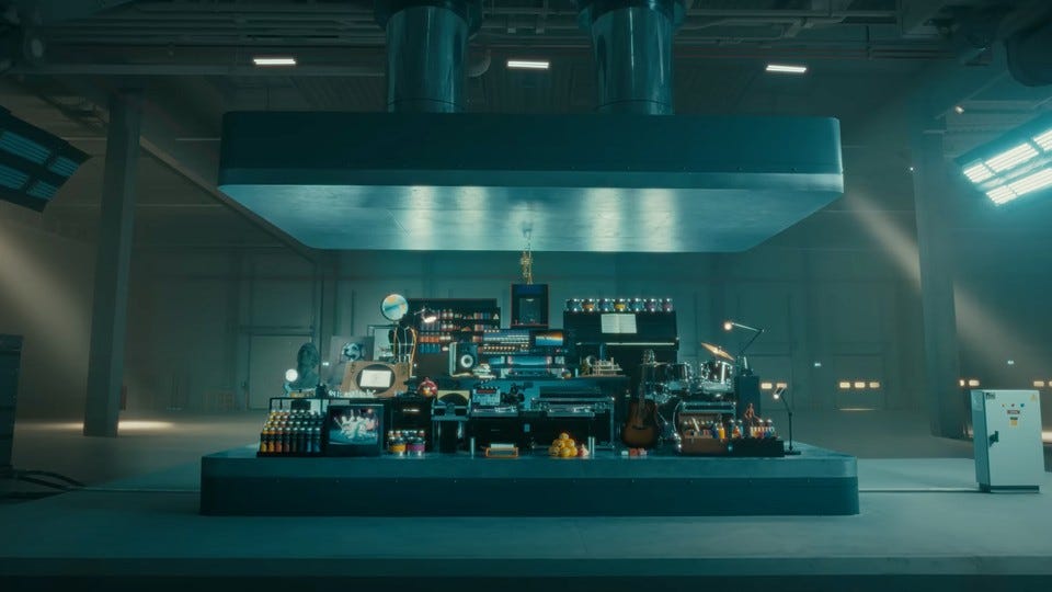 A large hydraulic press in a warehouse is poised above an assortment of cultural items, including a guitar and a piano.