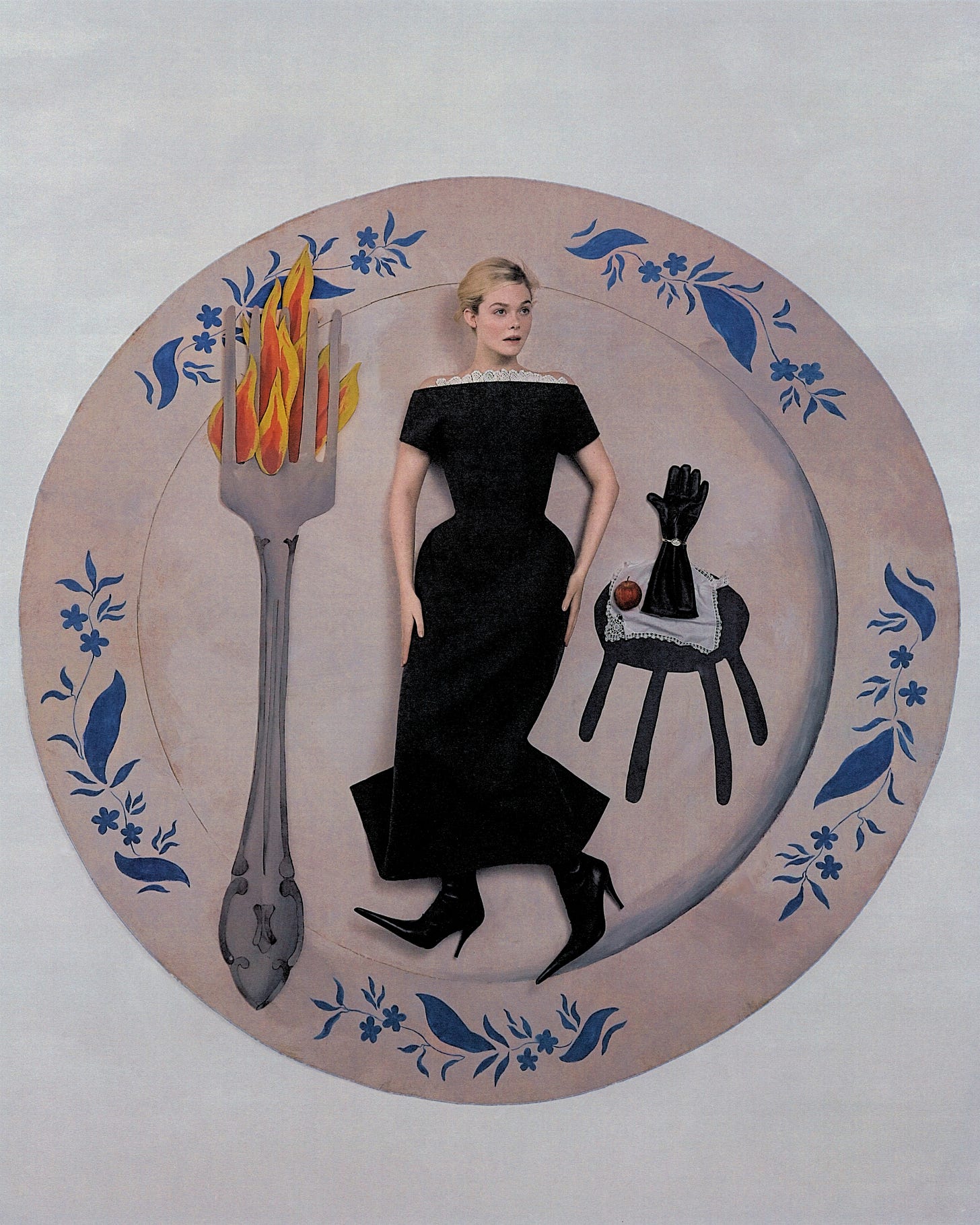 Elle Fanning shot by Szilveszter Mak&amp;amp;oacute;. Fanning is lying flat on a life-size cardboard cutout of a dinner plate. She is wearing a black off-the-shoulder dress with black pointed-toe boots. On her left is a large cardboard cutout fork with flames, and on her right is a cardboard cutout side table with black glove and apple placed on top.
