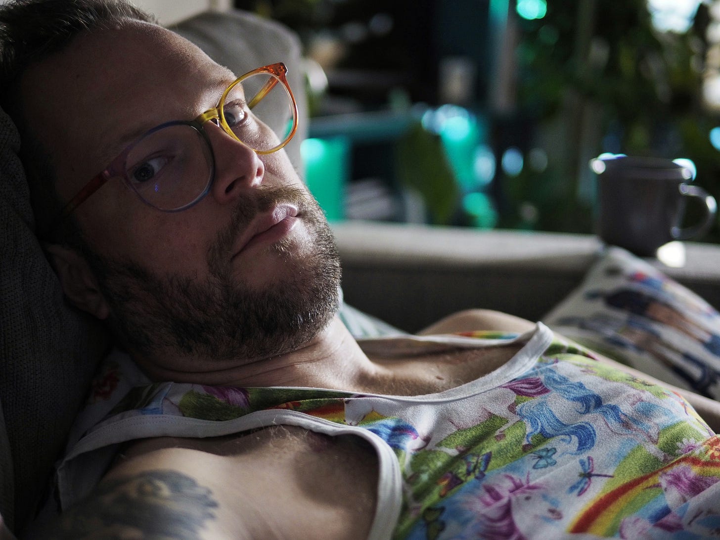 A photo of a white person with a beard reclined on a couch. they are looking at the camera through rainbow glasses. they wear a singlet with unicorns on it. 