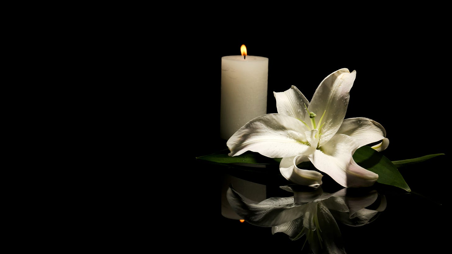A white lily rests beside a burning pillar candle on a glossy black surface, with their reflections visible against a dark background.