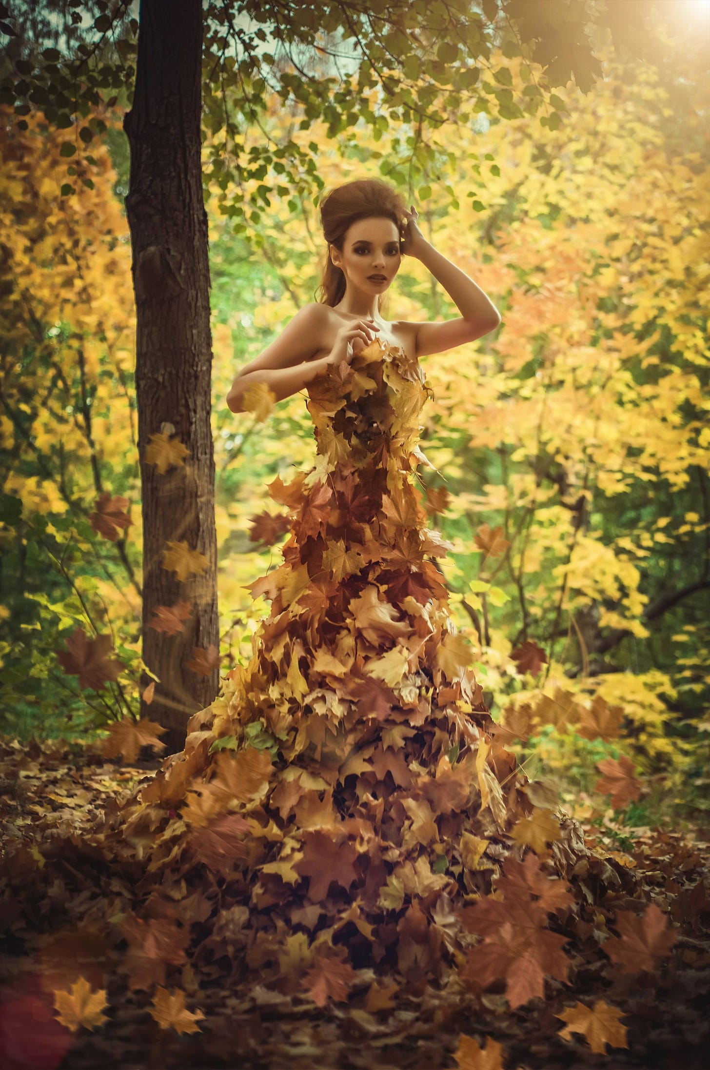 A woman stands in a sun-dappled forest, surrounded by trees with some green but mostly yellow leaves. Her brown hair is swept up and back, and she wears a strapless dress formed of numerous yellow and browning leaves. A woman stands in a sun-dappled forest, surrounded by trees with some green but mostly yellow leaves. Her brown hair is swept up and back, and she wears a strapless dress formed of numerous yellow and browning leaves.