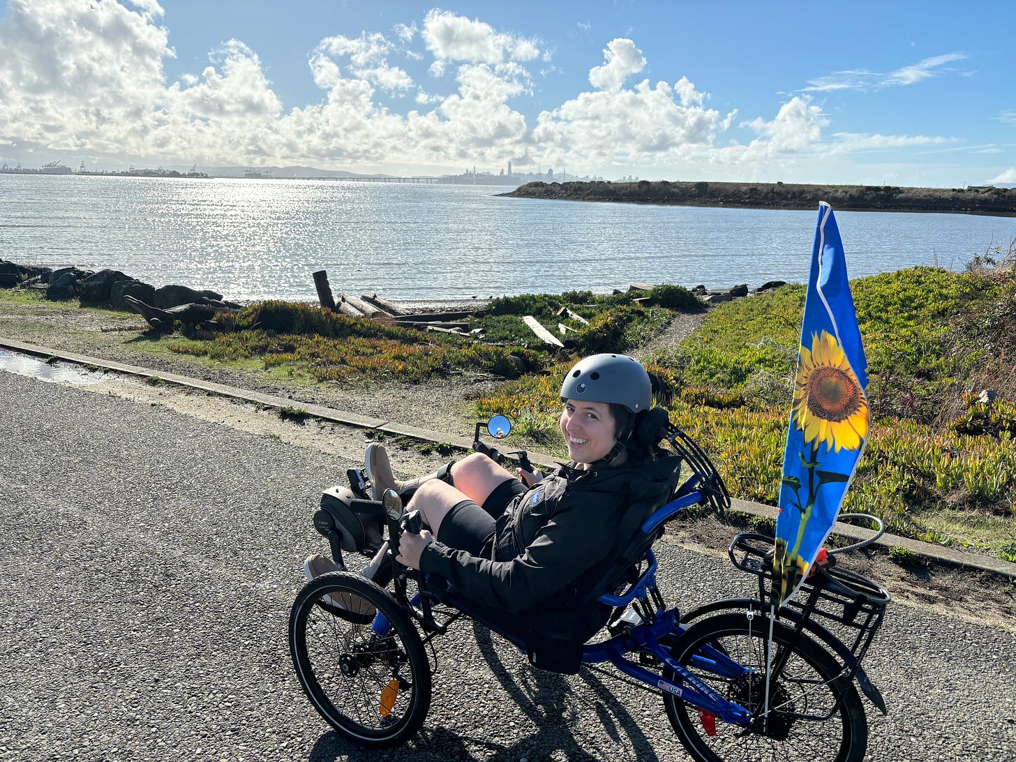 Image of Sonya Rio-Glick seated in a blue recumbent trike, wearing a helmet and smiling at the camera. The trike is parked along a cycling path and the sprawling waters of San Francisco Bay are in the background against a bright blue sky.