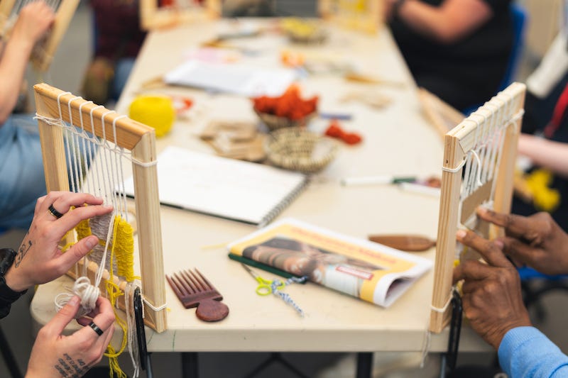 Two pairs of hands doing tapestry weaving on small wooden frames perched on the corners of a table