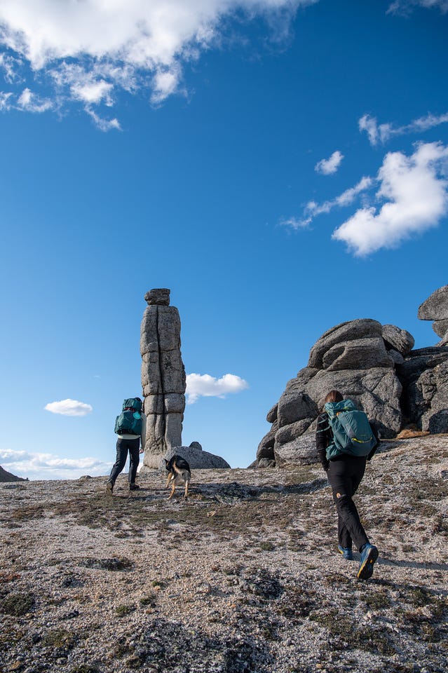 Two hikers with packs and a dog walk across pale, lichen-covered rock toward a tall, weathered granite pillar and a cluster of blocky outcrops under a blue sky.