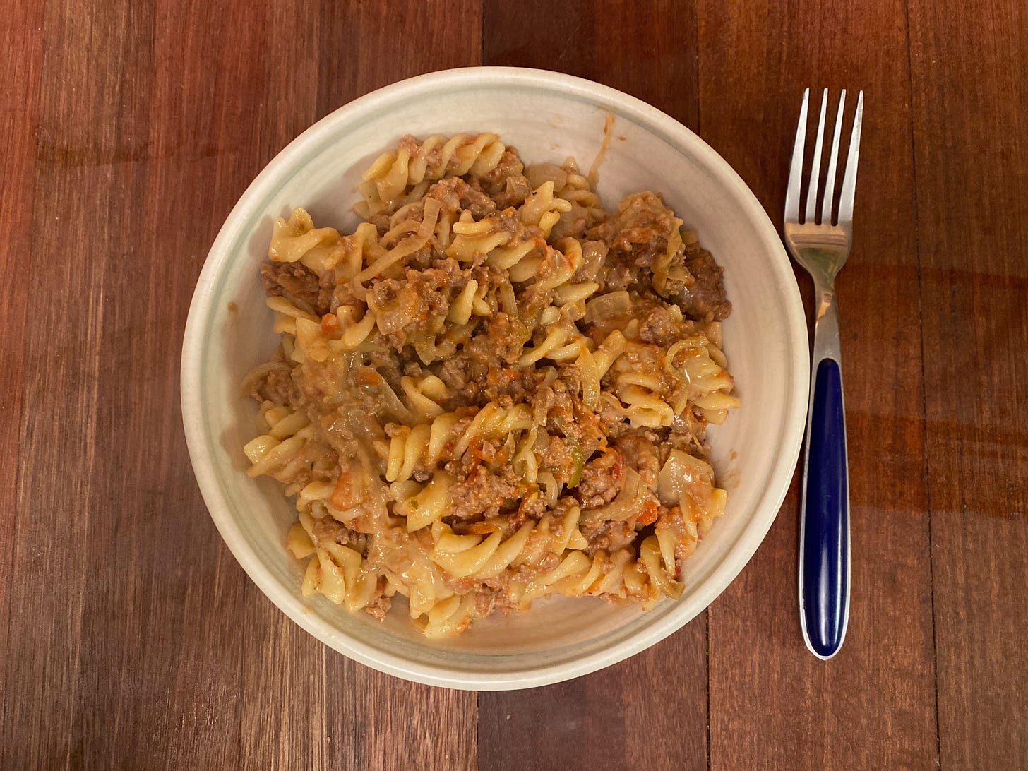 A white ceramic bowl filled with fusilli pasta tossed with a lamb tomato sauce. A fork sits on the wooden counter next to the bowl.