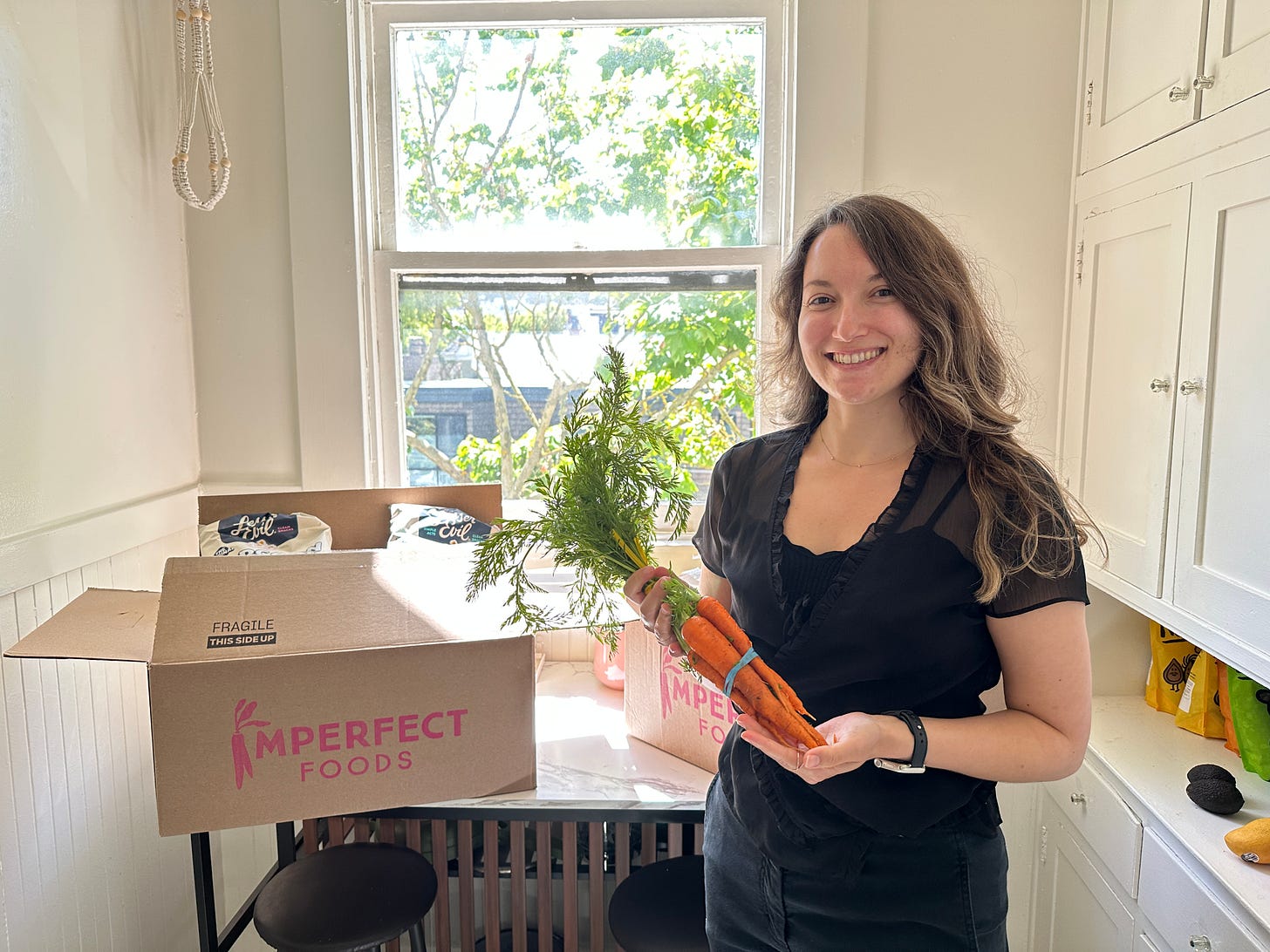 Photo of Amy Suto smiling in a sunny kitchen, holding a bunch of fresh carrots. Behind her are open boxes of Imperfect Foods produce. A candid look at Amy’s lifestyle and recommendation for healthy, sustainable grocery delivery for busy writers.